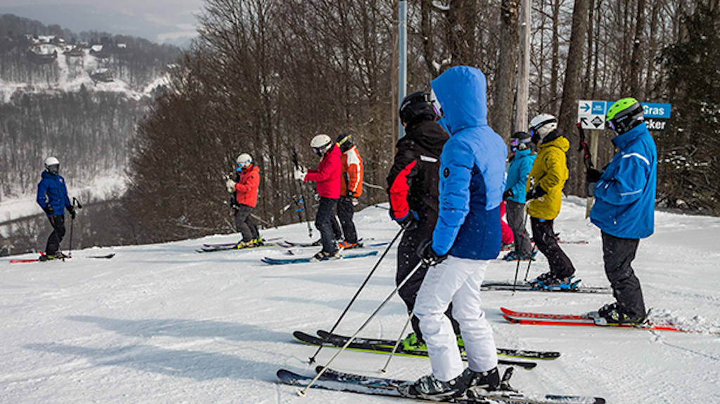 A group of adult skiers stand at the top of a hill and listen to their ski coach give them directions.